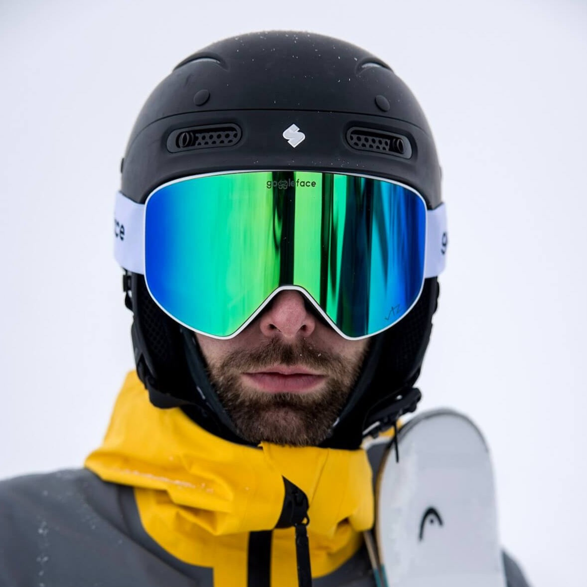 A person wearing a black helmet and Goggleface stands outdoors in the snow. Dressed in a yellow and gray winter jacket, they hold a Straight Line | White snowboard in their left hand, with a serious expression visible through the goggles.